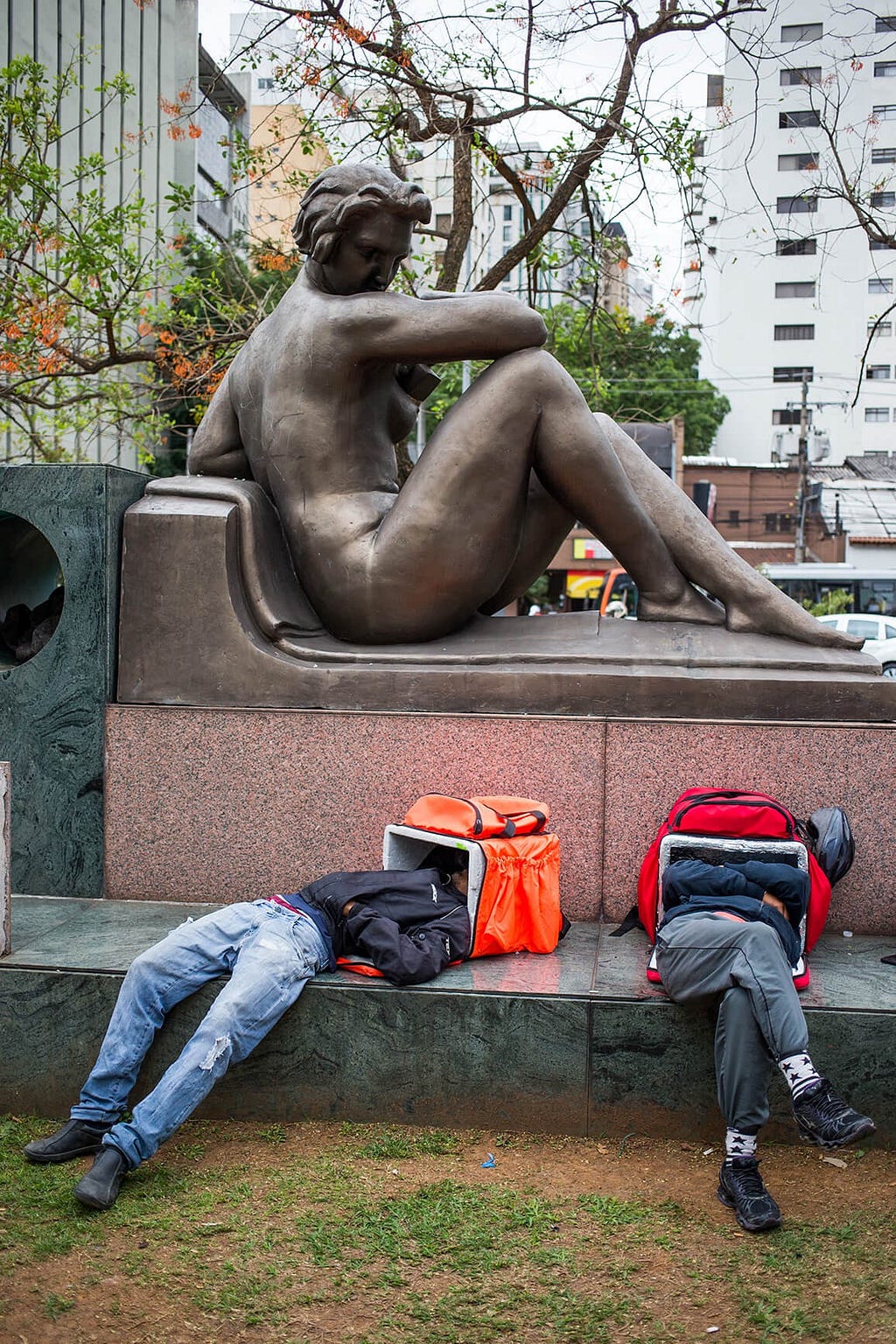 Dois homens estão deitados abaixo de um monumento de bronze, ambos estao com a cabeça enfiada dentro de uma mochila termica.