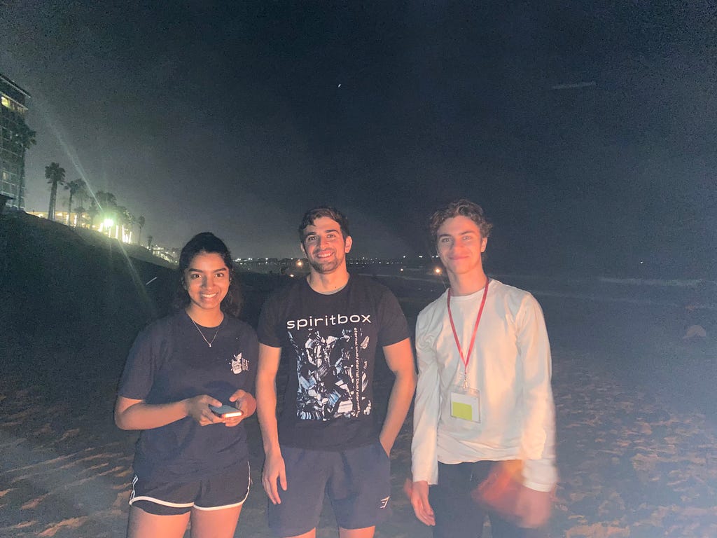 Three friends smiling on the San Diego beach together at night time.