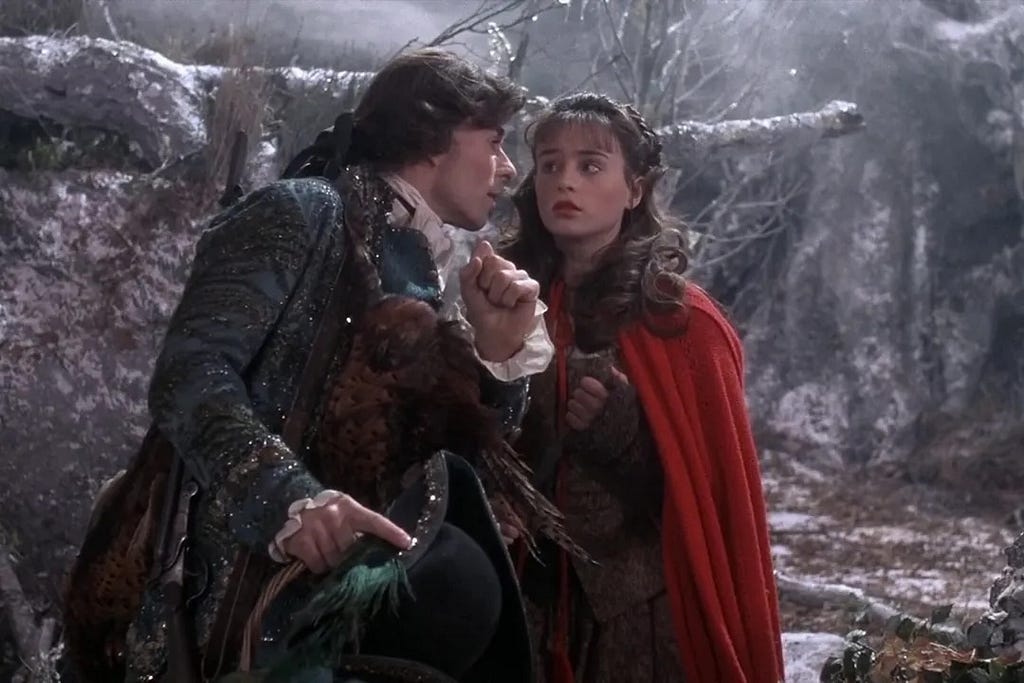In a snowy woods a  dark-haired girl in a red cloak — a ‘little red riding hood’ character — is being propositioned by a dashing man in 18th-century dress.