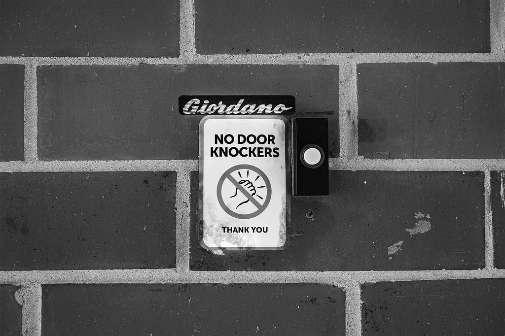 Doorbell with ‘no door knockers’ sign next to it. Above doorbell, ‘Giordano’ sticker.