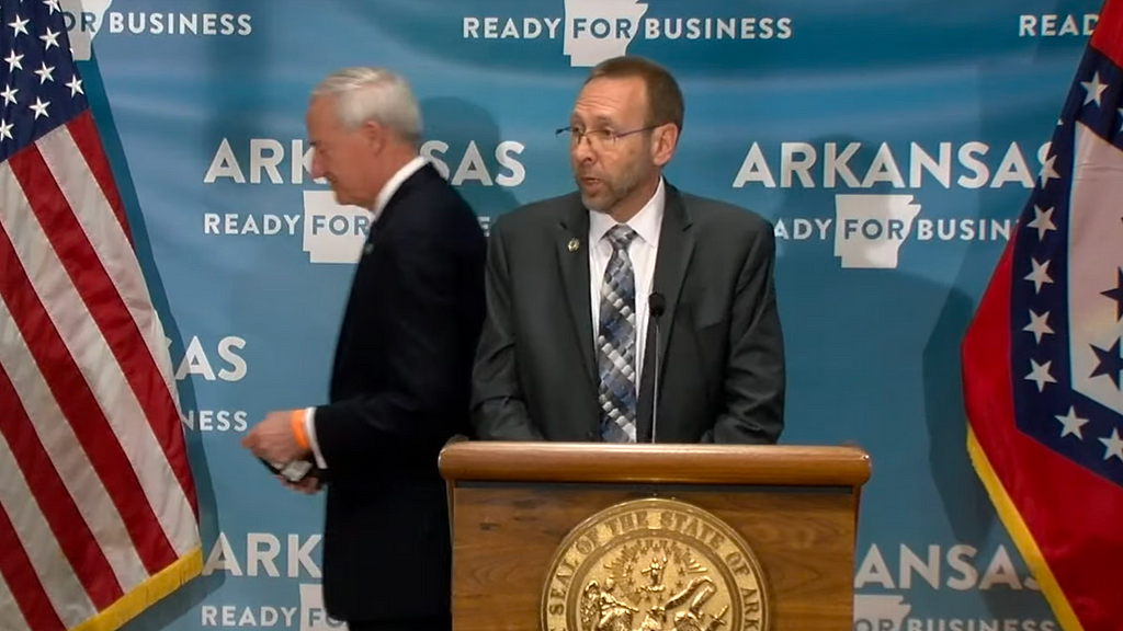 Arkansas Secretary of Health Dr. Nathaniel Smith speaks at a podium as Governor Asa Hutchinson passes behind him.