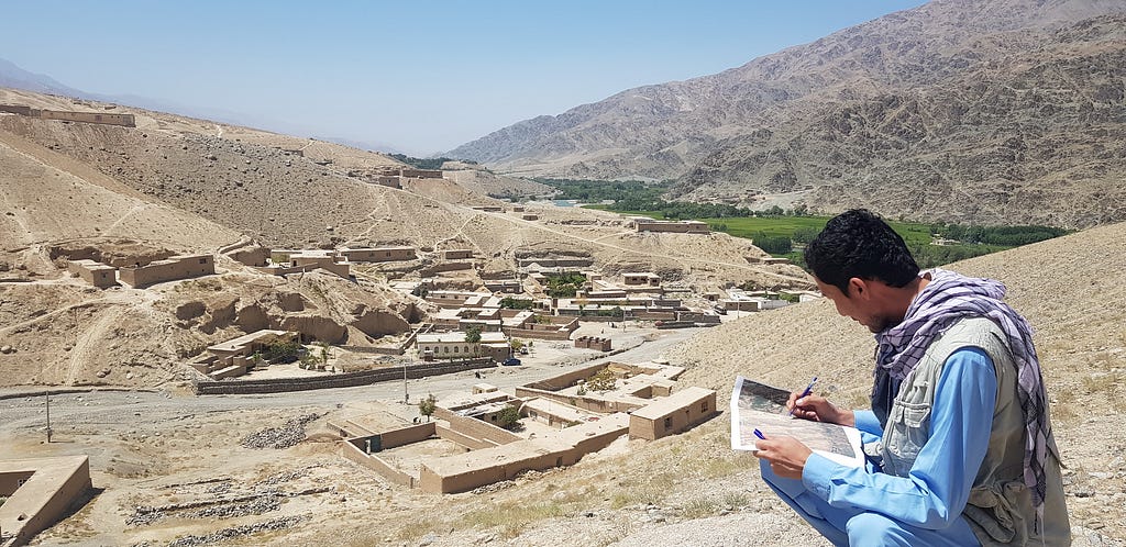 A man crouches and records notes on a map. In the background, there is a village at the foothills of two mountains.