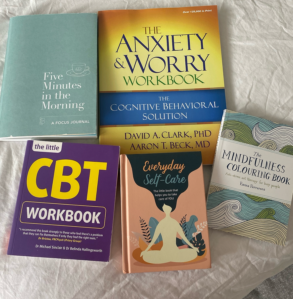 Image of five books on a bed with white bed sheets. The top left book is blue and titled ‘five minutes in the morning.’ It is a journal. The second book is a CBT and worry workbook- the cognitive behavioural solution. The third is a mindfulness colouring book and it has an ocean template on the cover. The third is a book titled ‘Everyday self care’. The last is a purple CBT workbook.