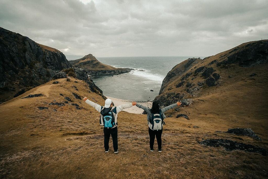 Two travelers stand at the top of a cliff and overlook the sea with their arms outstretched.