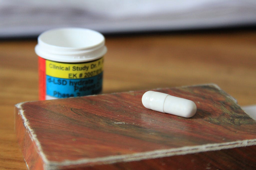 A white pill on top of a wodden box with a prescription bottle behind it. Photo courtesy of the Multidisciplinary Association for Psychedelic Studies