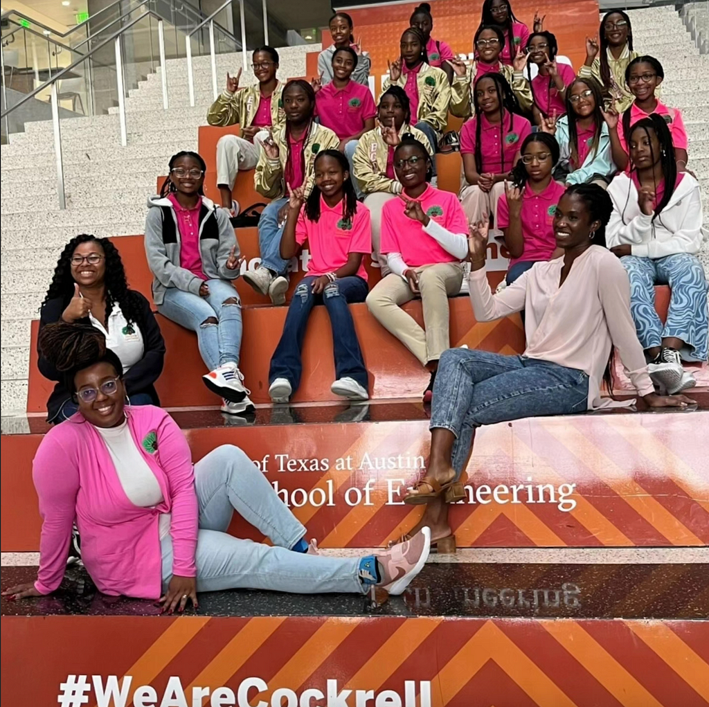 Group of Black Girls Do Engineer members and leadership sitting on the steps at the University of Texas at Austin School of Engineering. The stairs are shades of orange with light and dark orange strips. One of the steps has The University of Texas Austin School of Engineering on it and the another steps has #We are Cockrell on it. The girls are holding up their pinky and pointer fingers representing the horns for the University of Texas mascot. All of the girls are wearing pink with a BGDE logo