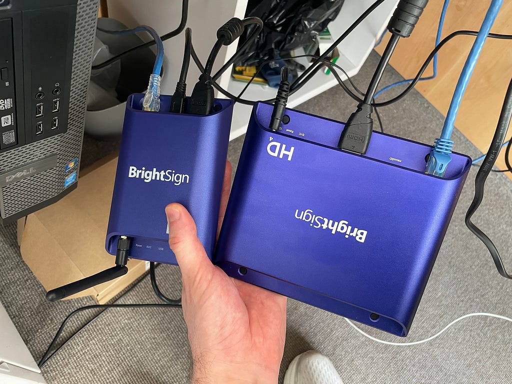 Two purple BrightSign players held in a hand, with cables running out of them.