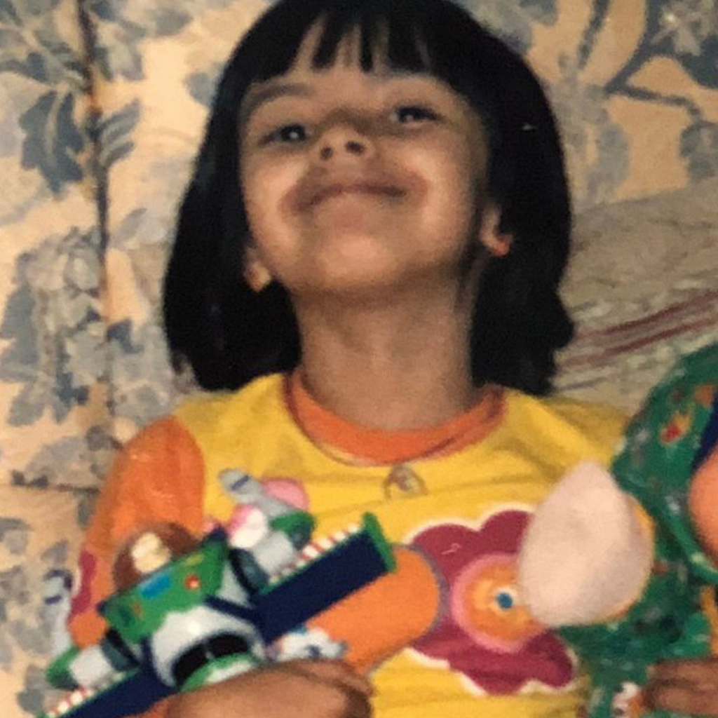 Uma foto minha de quando eu era criança. Estou sentada no sofá, sorrindo para a câmera, e segurando dois brinquedos, um em cada mão.