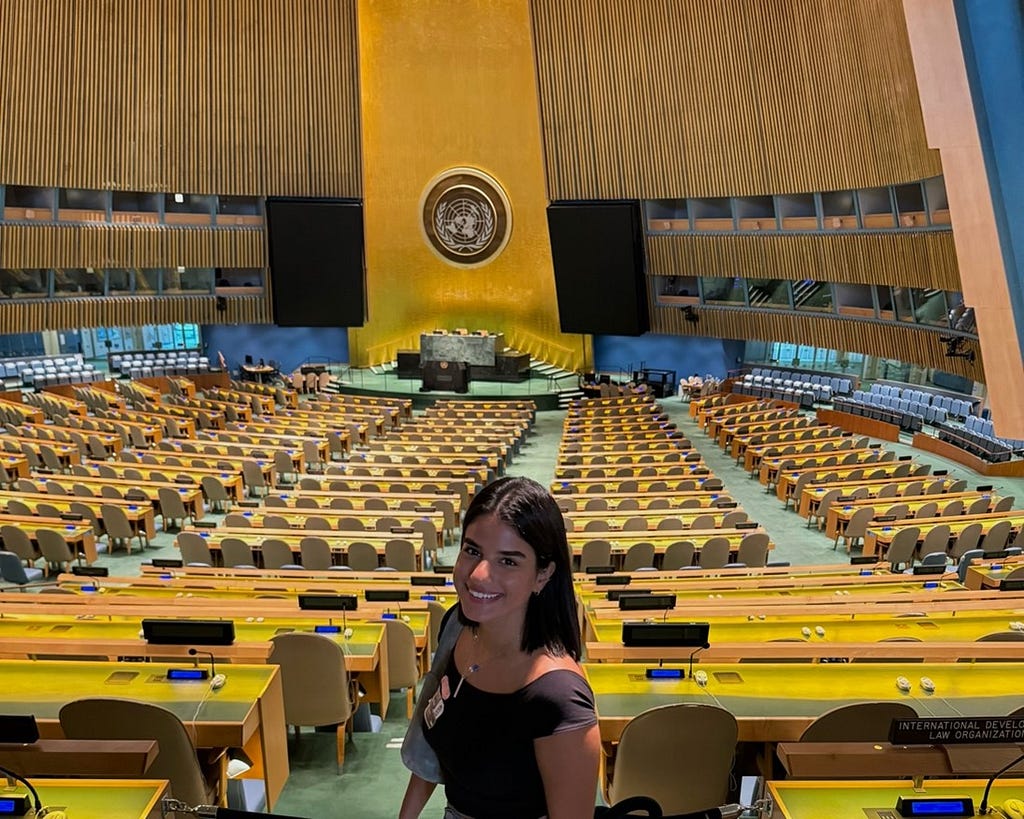 Sarah wearing black and smiling in front of the UN General Assembly: a yellow and blue room with a lot of yellow chairs.