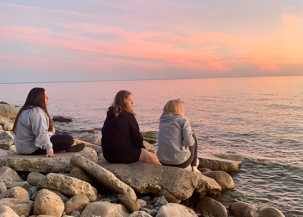 3 teenage girls sitting on scattered rocks along a lake, watching a colorful sunset.