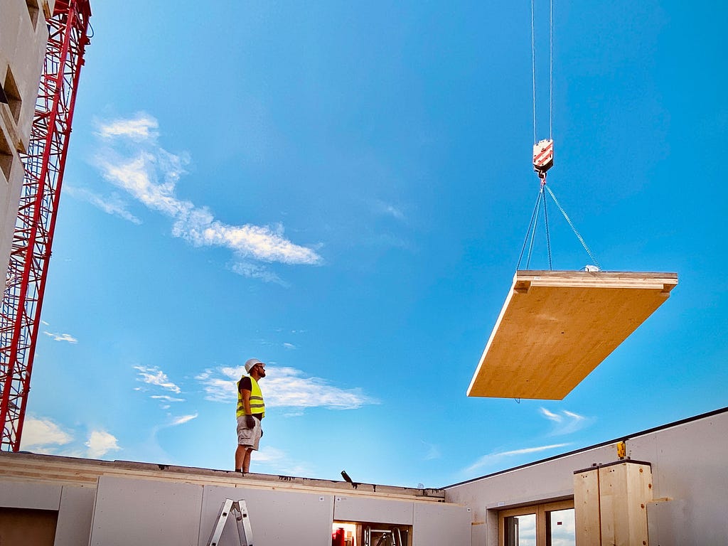 Man in neon yellow vest standing on wall unfinished apartment, looking at a wooden slab lifted by a red crane that is hanging in the air.