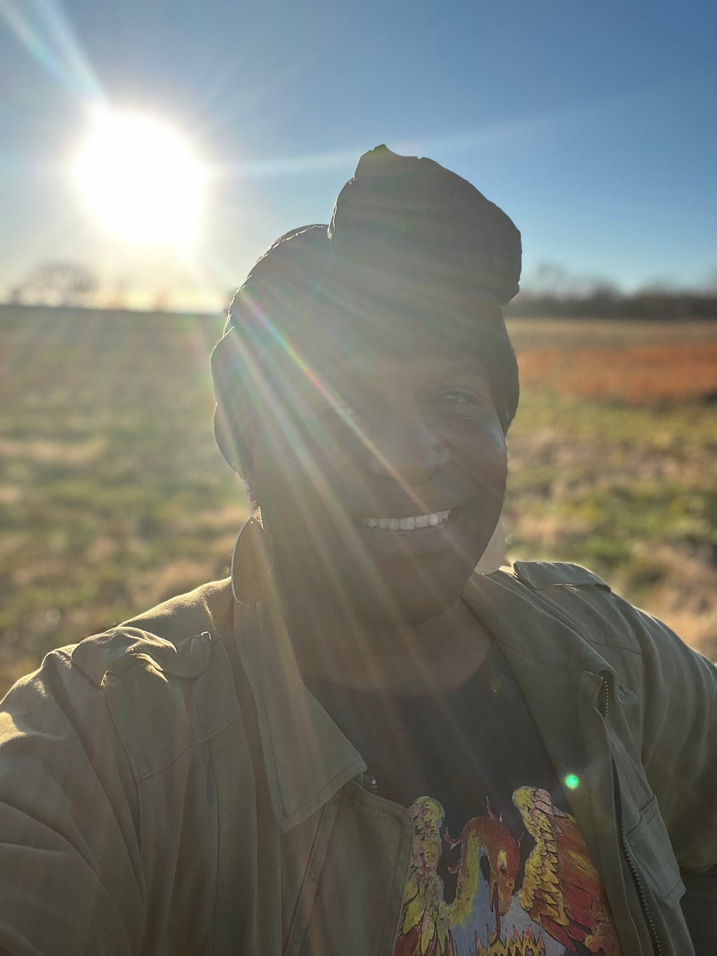 Black trans human standing in a field with the sun on their back. the human is smiling. assured that what will be, will be.