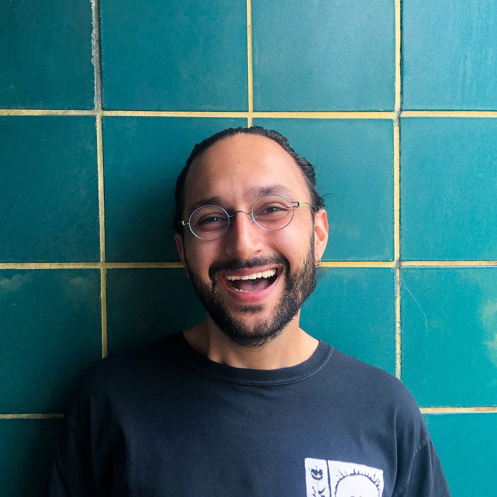 Bhavik stands smiling widely in front of a green tiled wall. He has a full, neat beard , colorful but subtle glasses, and his long hair is up in a tight high bun. He is wearing his favorite t-shirt, which has a stylized image of a sun on it.
