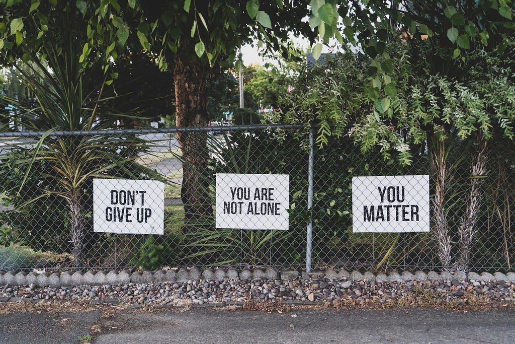 Three signs that say “Don’t give up”, “You are not alone”, and “you matter” are written in black ink on white backgrounds, hanging off a wired fence enclosing forestry.