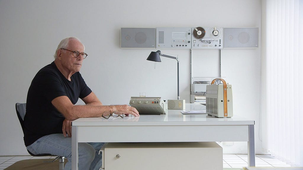 Still image of designer Dieter Rams sitting at a desk, surrounded by objects he has designed.