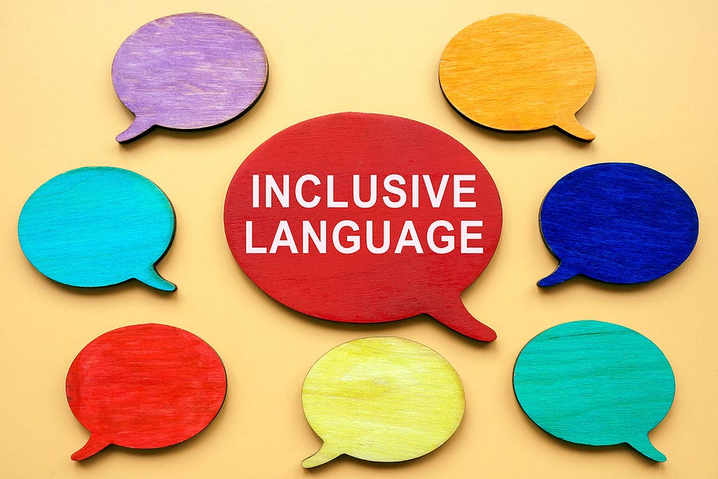 An image features a yellow background with eight colorful wooden speech bubbles arranged in a circular pattern. The speech bubbles are painted in various colors: purple, orange, blue, teal, green, yellow, red, and another red one in the center. The central red speech bubble has the words ‘INCLUSIVE LANGUAGE’ written in white capital letters. The arrangement and bright colors create a vibrant and engaging visual emphasizing the importance of inclusive language.