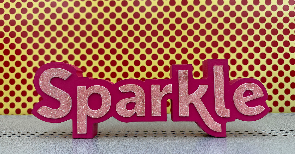 Pink sign that says “Sparkle” against a yellow backdrop with pink polkadots.