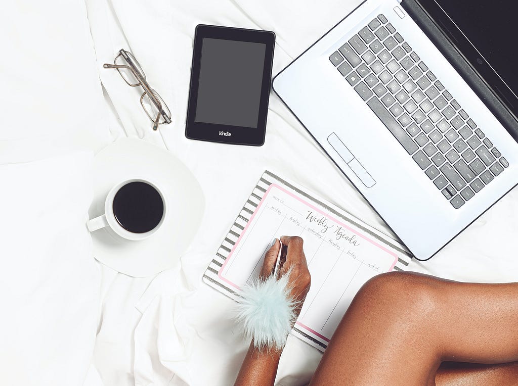 A pair of glasses, mug of coffee, person writing with decorative pen on a calendar, Amazon Kindle, and laptop.