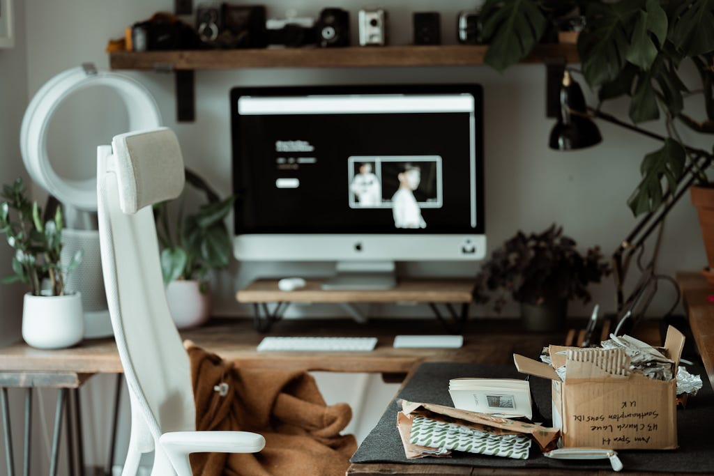 An office space is viewed. A white chair, desktop with an online store website on display and packaging materials and office supplies scattered around.