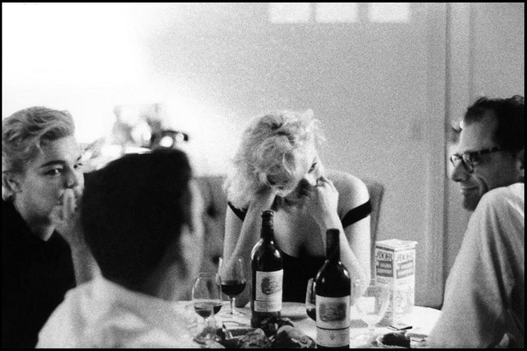 Black and white candid photograph of a dinner with four famous people deeply engaged in conversation around a small kitchen table: Arthur Miller, Marylin Monroe, Simone de Beauvoir, and actor Yves Montand.