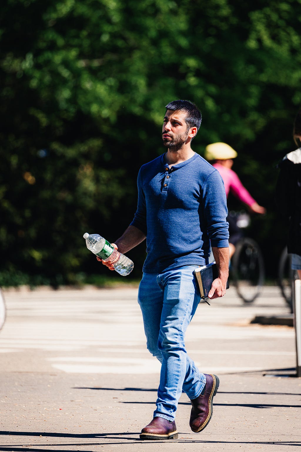 Man in jeans with a water bottle walking in front of green trees