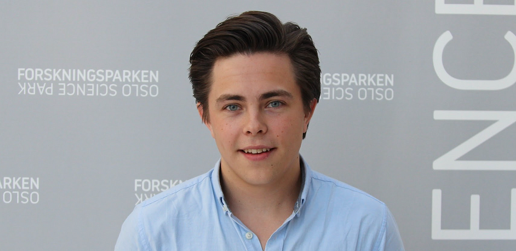 Picture of the author wearing a blue shirt standing in front of a grey background