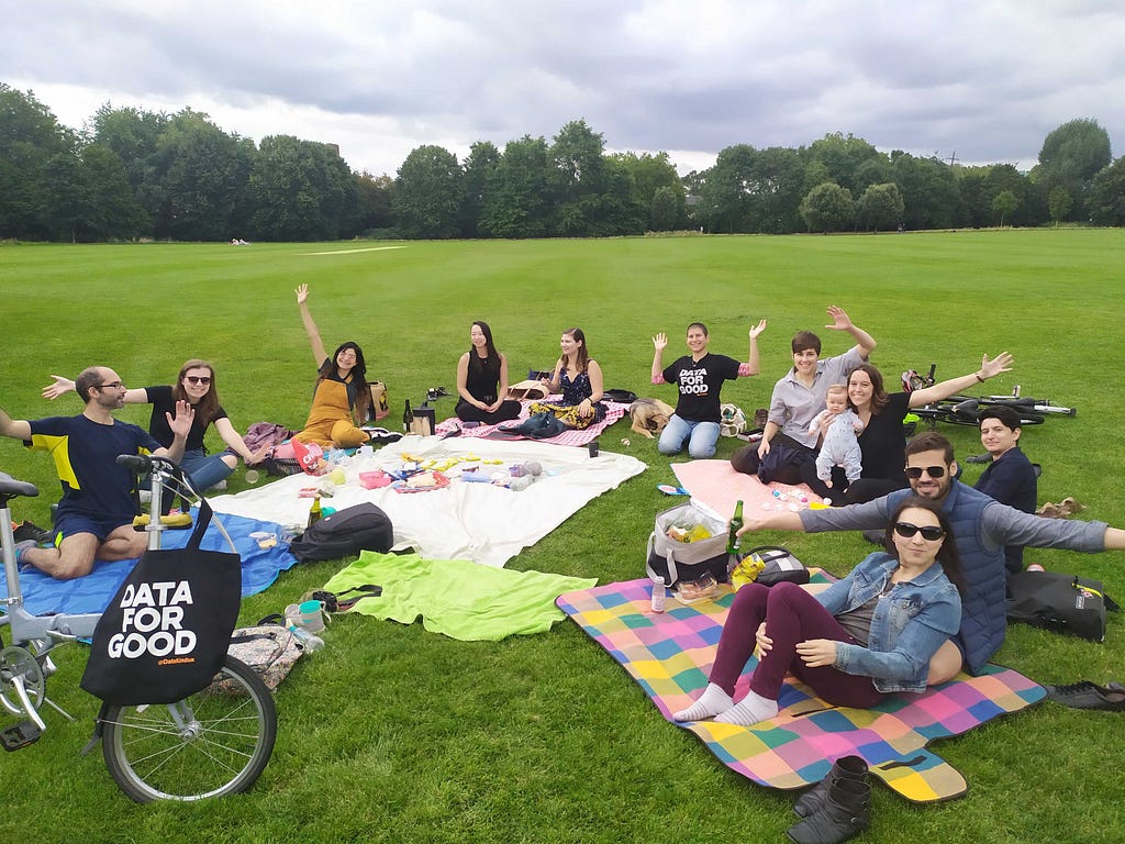A group of adults sat outside in a London park for a picnic, waving and smiling, with a grey sky overhead