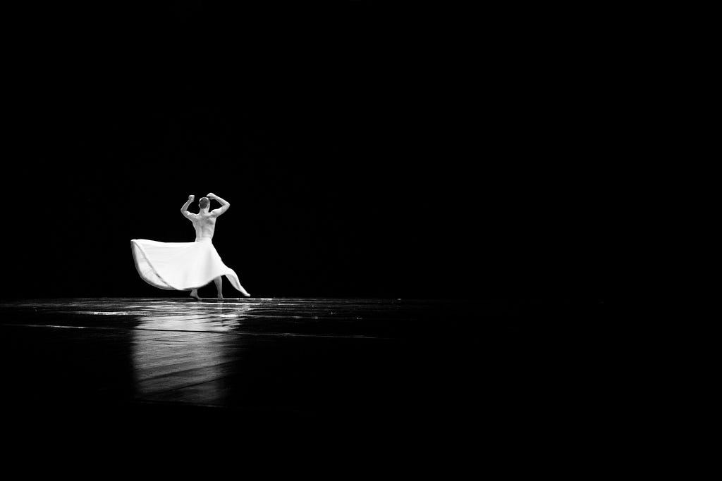 A dancer in a white dress dancing alone on a stage steeped in darkness.