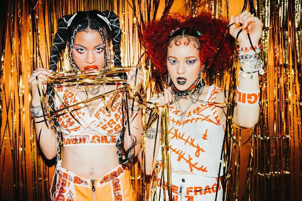 Guitarist/vocalist Amy Love and bassist Georgia South, two young Black women, pose in front of an orange Mylar curtain