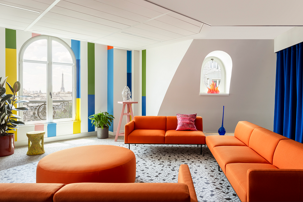 A cosy lounge with orange couches, lighten by a window where we can see the Eiffel Tower