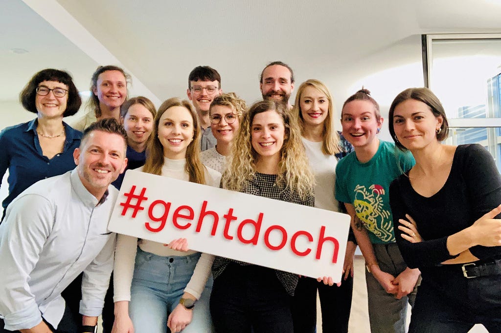 12 Personen jüngeren und mittleren Alters mit heller Haut und informeller Kleidung stehen als Gruppe und schauen direkt in die Kamera – alle lächeln, eine Frau ganz vorn hält ein Schild mit dem Hashtag #gehtdoch hoch