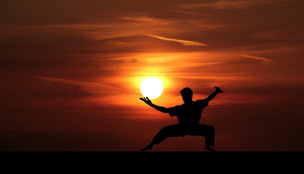A martial artist in a low stance silhouetted against a deep orange sunset, holding the sun between his hands as if in balance; an image symbolising self-mastery, perception and inner leadership.