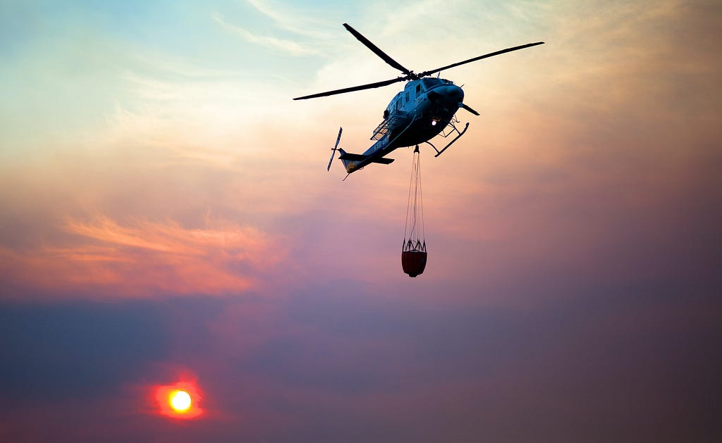 A firefighting helicopter is seen flying overhead with its bucket at sunset. The sun is a deep orange-red as it sets in sky filled heavily with smoke.