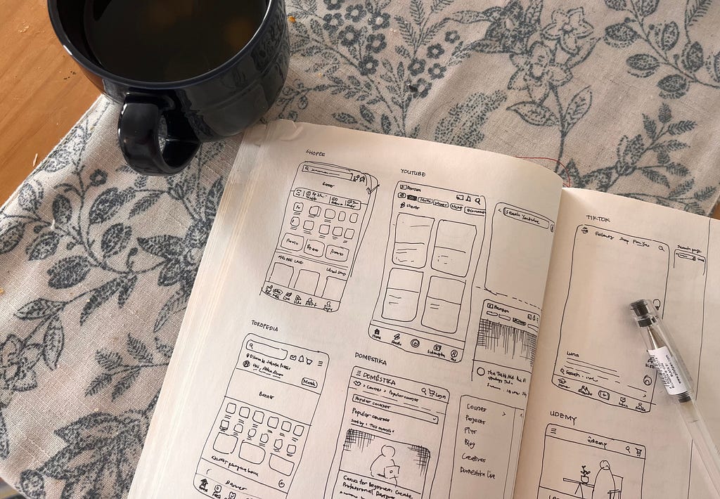 A cup of tea, a notebook with sketches of mobile UI, and a pen on top of a table runner.