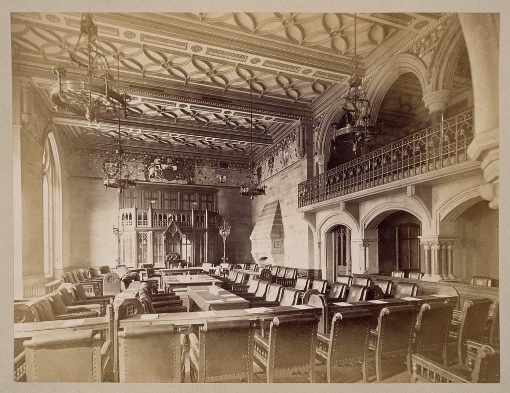 Elaborately decorated room with tall windows, a gallery, stone fireplace and arches. Lots of chairs facing the Mayor’s dais.