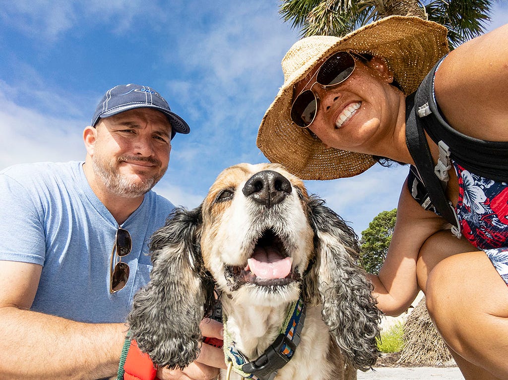 A man and lady in a sunny tropical location taking a photo with a dog