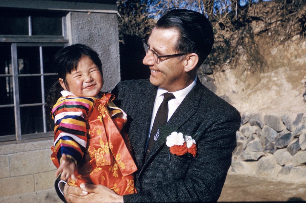 Rev. Swanson is wearing a suit and tie and carrying a Korean toddler in his arms. They are both smiling.
