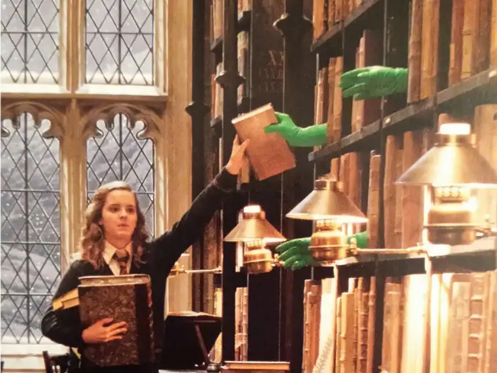 Hermione Granger (Harry Potter) segurando um livro grande com um dos braços enquanto estica a sua mão para o alto para buscar outro livro no alto de uma prateleira sendo entregue por mãos com luvas verdes para ser usado efeito especial.