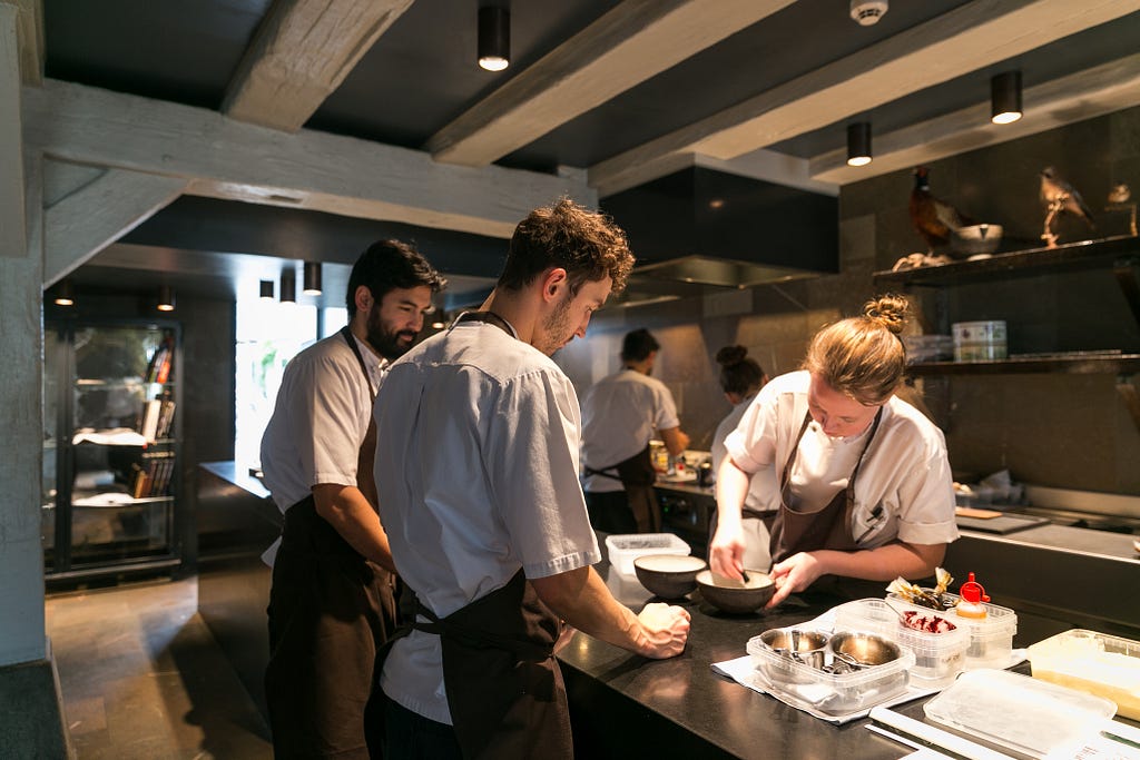 Three chefs working together in a modern restaurant kitchen, with another two chefs working at a different station in the background.