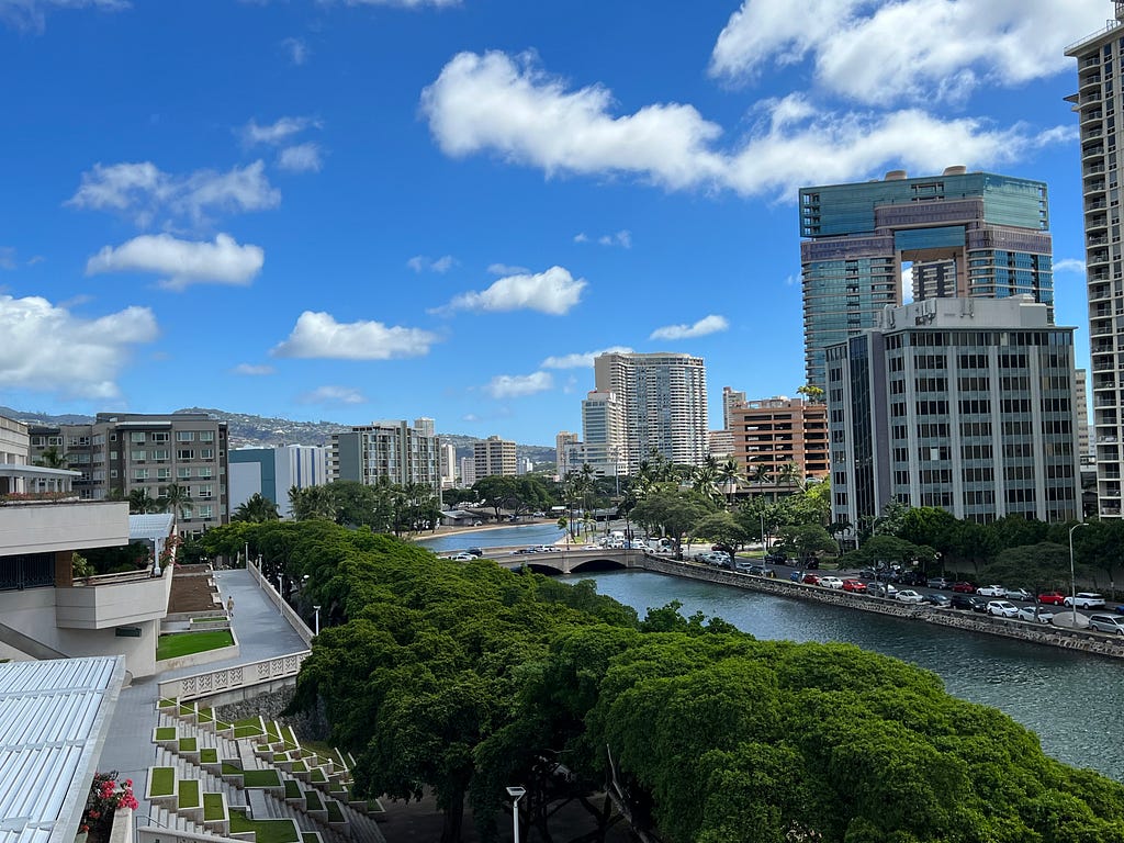 Photo of the view from a Honolulu hotel window; deep blue sky, tall buildings on the right, green trees and water in the foreground