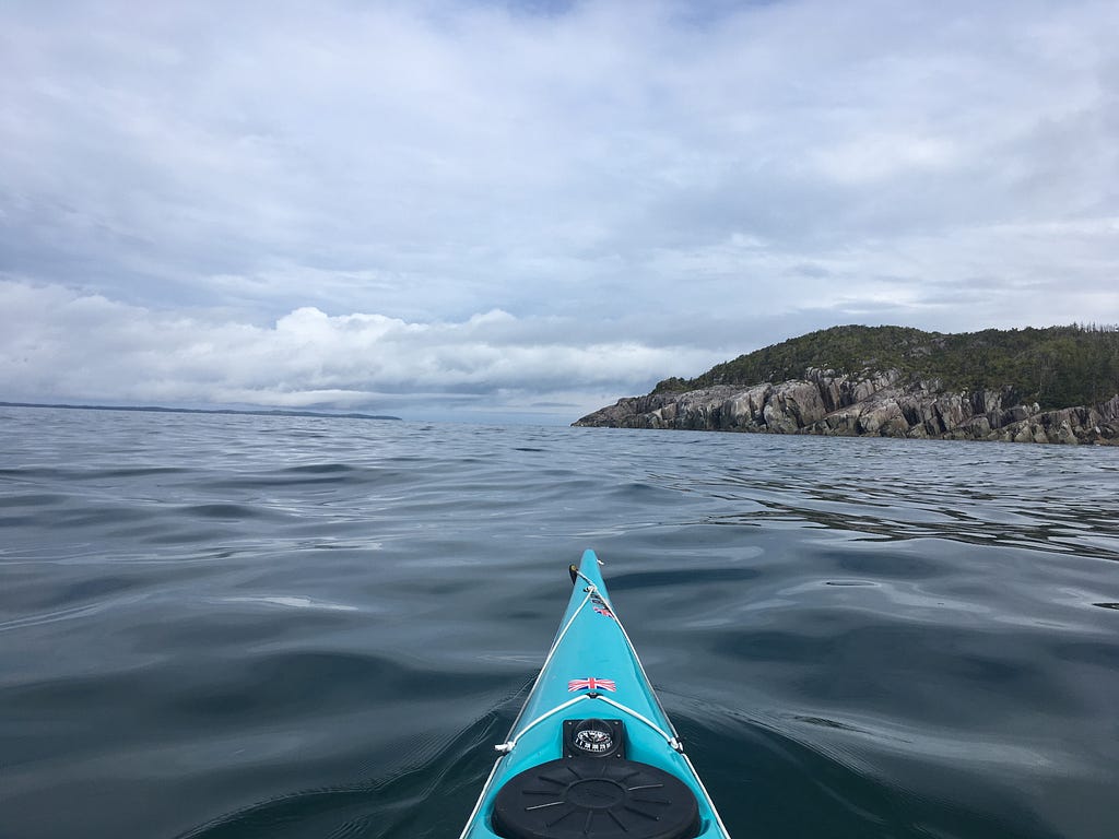 The nose of a kayak in the foreground with a nearby rocky bluff and a distant island in the background.