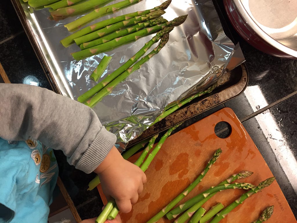 A small child puts asparagus on a baking sheet.