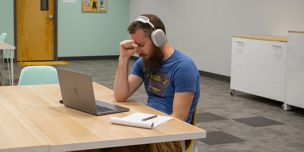 A man wearing headphones, sitting at a desk in front of a computer with his eyes closed in sadness