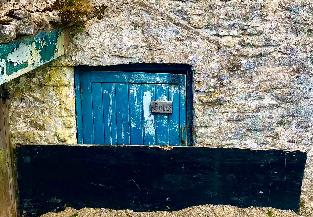 A small door nestled within a stone wall. There is a small sign which reads: “Pull”.