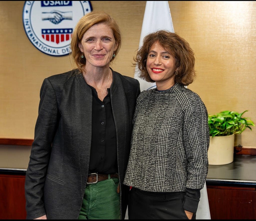 Two women stand next to each other in front of a USAID seal and flag.