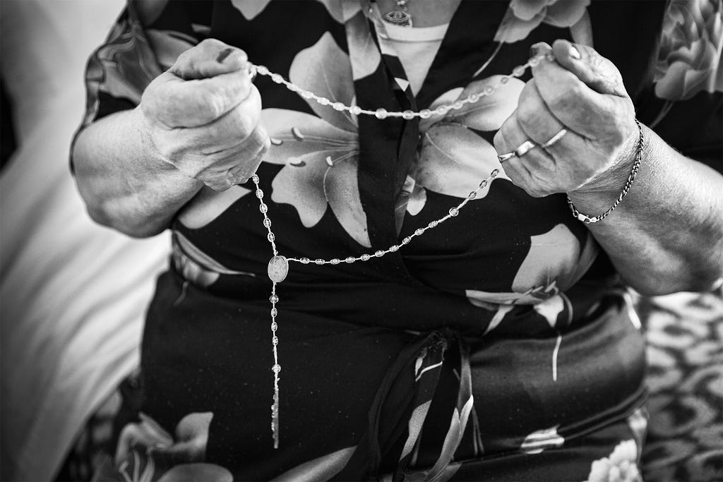 Stefani’s grandmother prepares to put her rosary on in the morning. She is holding it between both of her hands. Only her torso and hands are visible.
