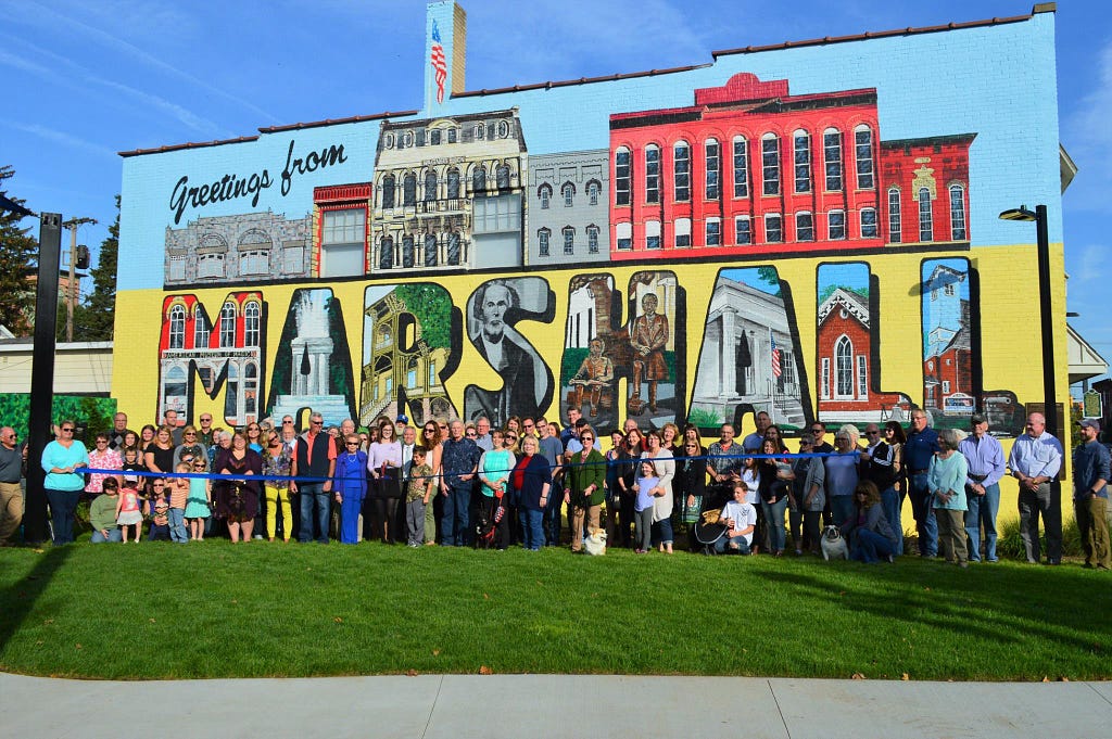 The ribbon cutting ceremony for Grand Street Park in Marshall, Michigan. Hundreds of community members stand behind a blue ribbon in the park in front of a large mural with the words, “Greetings from Marshall.”