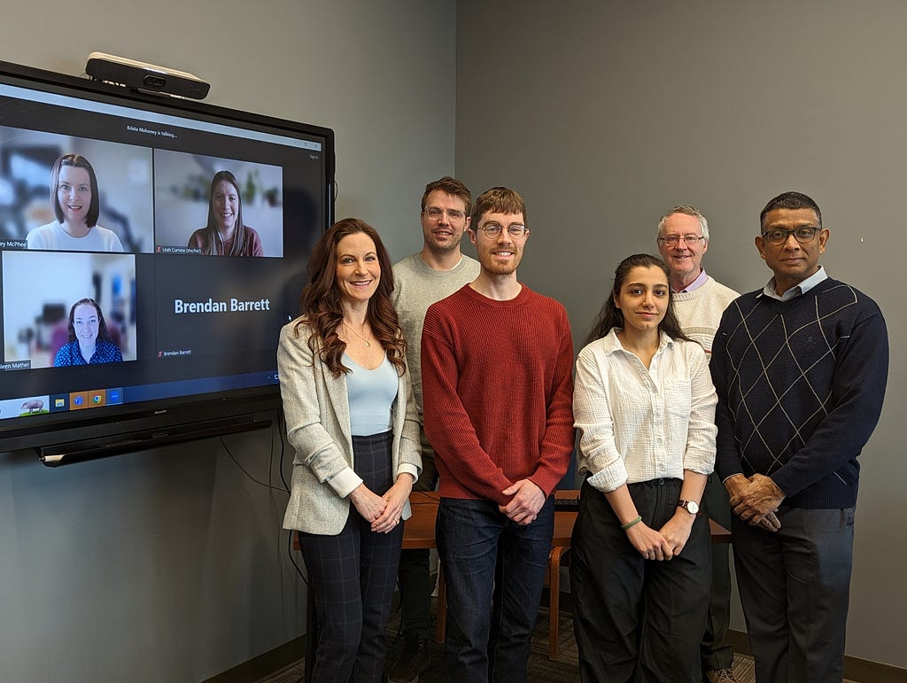 A group photo of nine people smiling. Three people are smiling on a tv screen from a virtual platform.