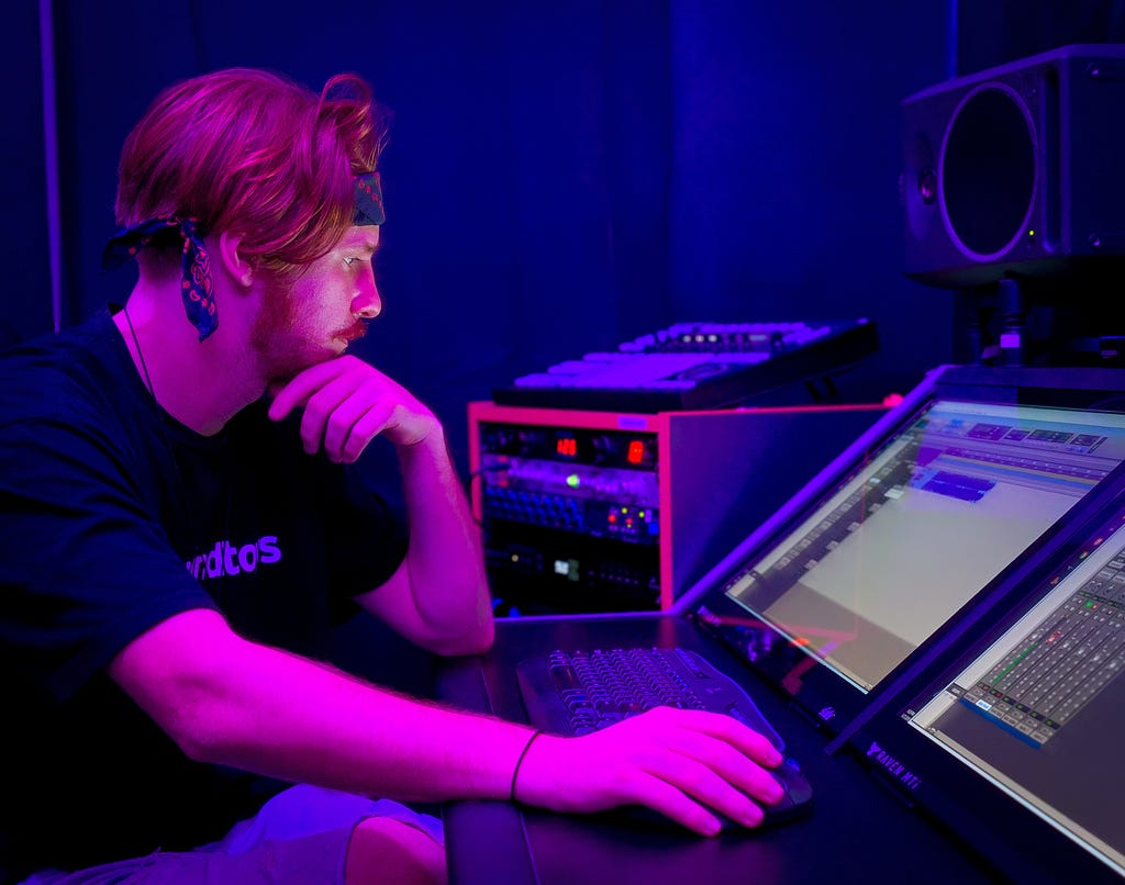 a person using a digital sound mixing program with a mouse and keyboard. There are a few pieces of sound equipment like a euro-rack and subwoofer near them.