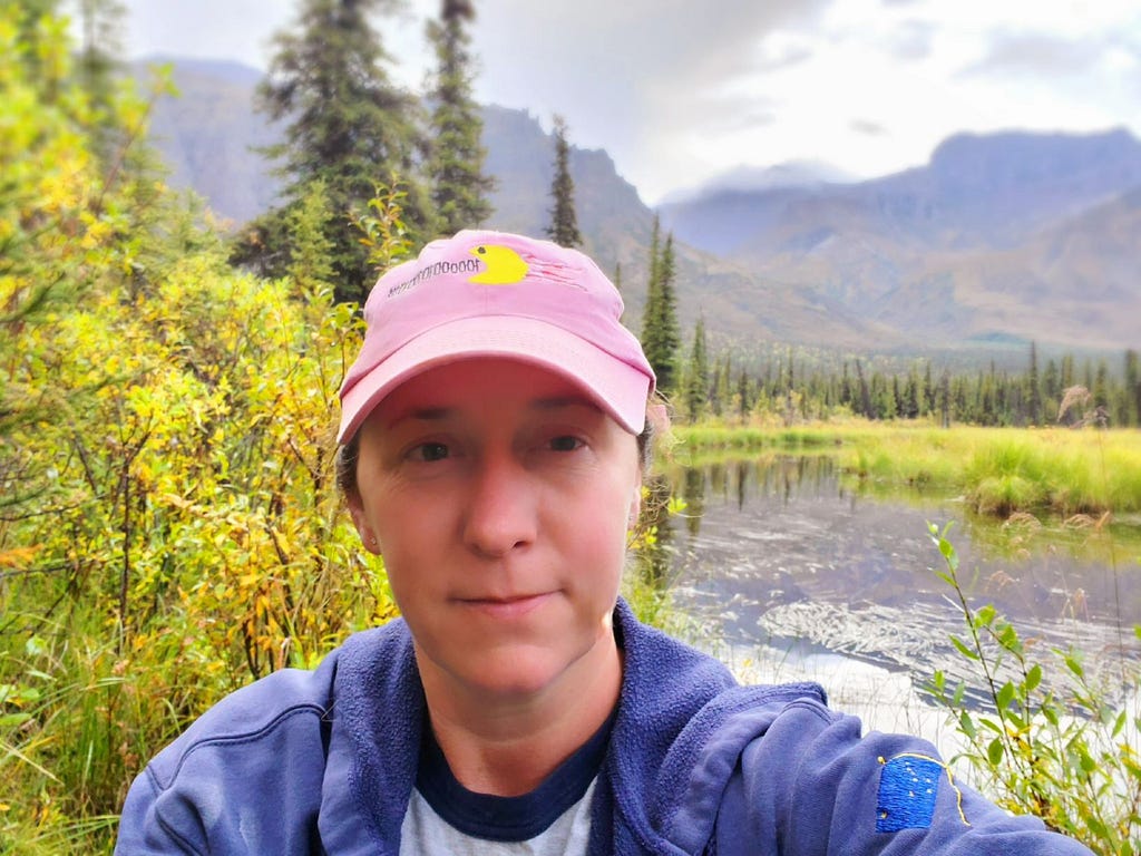 Woman wearing a pink baseball hat and blue hoodie in front of a small lake and mountains. spruce trees surround lake